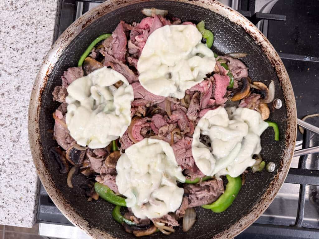 Sliced beef, green bell peppers, and onions in a frying pan, topped with melted cheese.