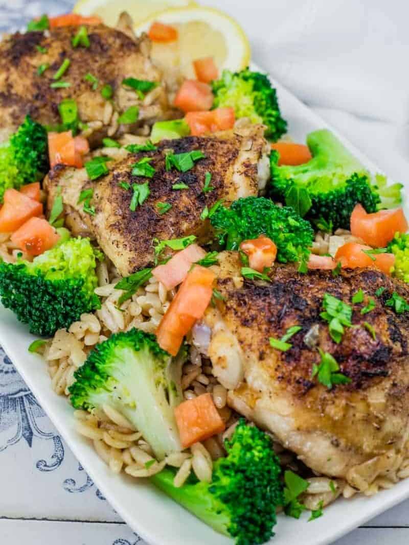 A Lemon Chicken Orzo Skillet with Broccoli dish featuring golden-brown seasoned chicken thighs on a bed of orzo pasta, garnished with fresh broccoli florets, diced tomatoes, chopped parsley, and lemon slices in the background.