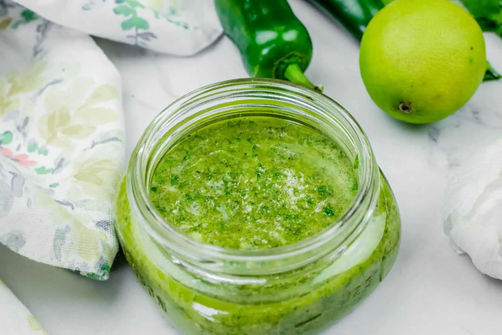 An open glass jar filled with Lime-Garlic Cilantro Marinade sits on a white surface next to a green chili, a lime, a garlic bulb, and a patterned cloth.