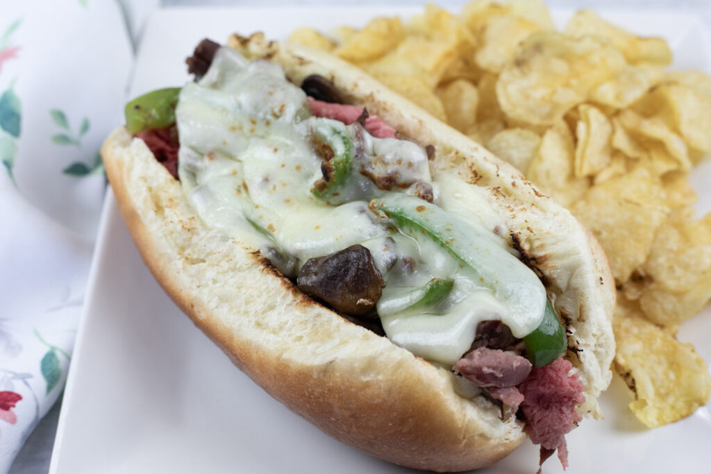 A sandwich with melted cheese, green peppers, and meat in a bun, served with a side of potato chips on a white plate.