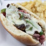 A sandwich with melted cheese, green peppers, and meat in a bun, served with a side of potato chips on a white plate.