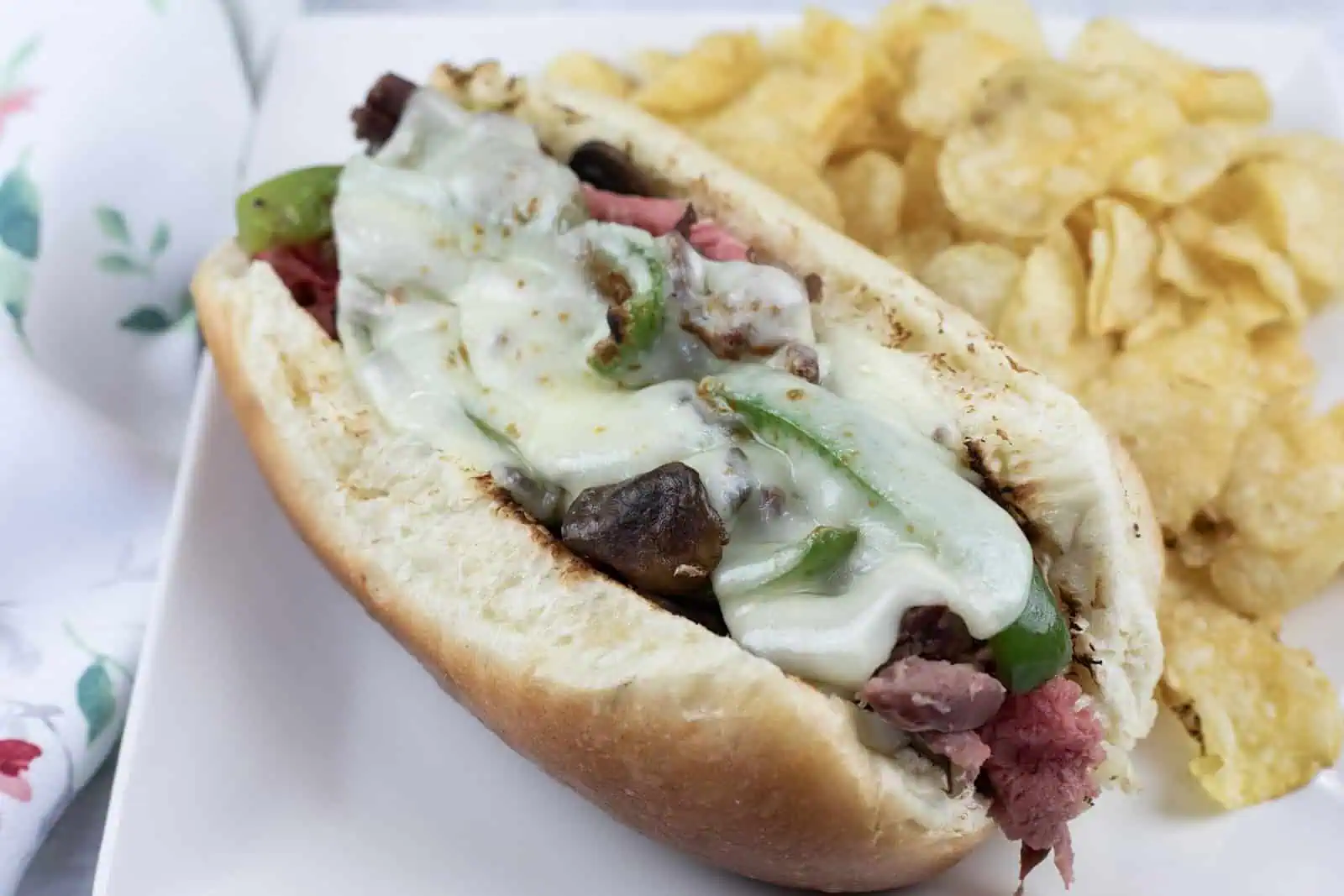 A sandwich with melted cheese, green peppers, and meat in a bun, served with a side of potato chips on a white plate.