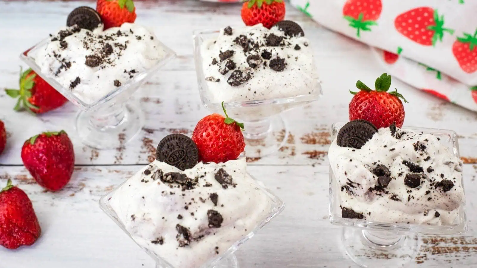 Four glass dessert cups filled with cookies and cream mousse, each topped with a whole strawberry and a mini chocolate cookie, with strawberries scattered on a white wooden surface.