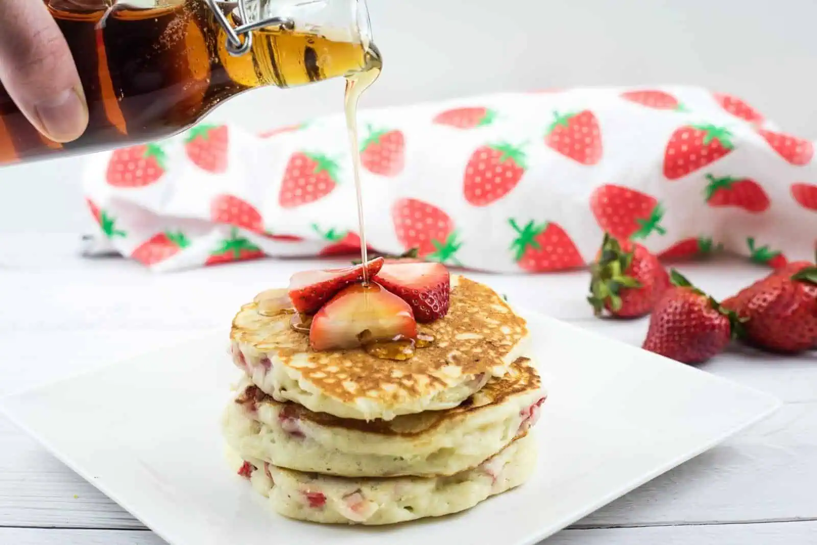 A stack of pancakes with sliced strawberries on top is being drizzled with syrup, with whole strawberries and a strawberry-patterned cloth in the background.