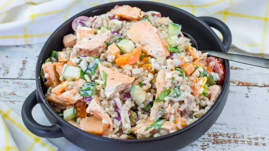 A black bowl filled with a mixed salad containing chunks of salmon, rice, cucumbers, carrots, onions, and herbs, with a fork on the side.