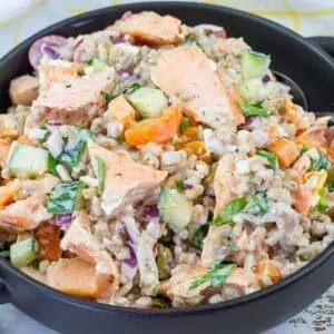 A black bowl filled with a mixed salad containing chunks of salmon, rice, cucumbers, carrots, onions, and herbs, with a fork on the side.