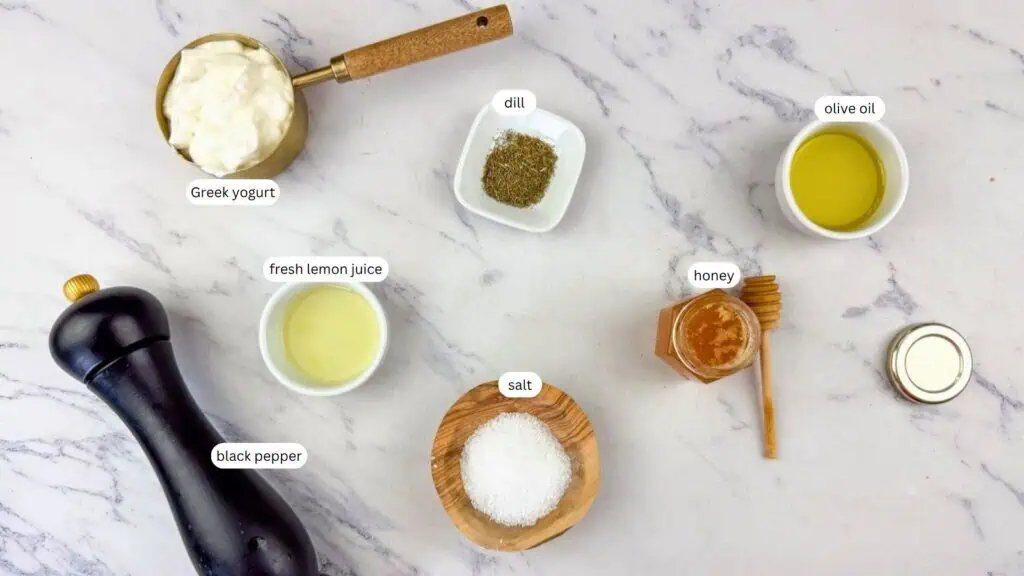 Ingredients on a marble surface: Greek yogurt, dried dill, olive oil, fresh lemon juice, honey, salt, and a pepper grinder labeled black pepper.