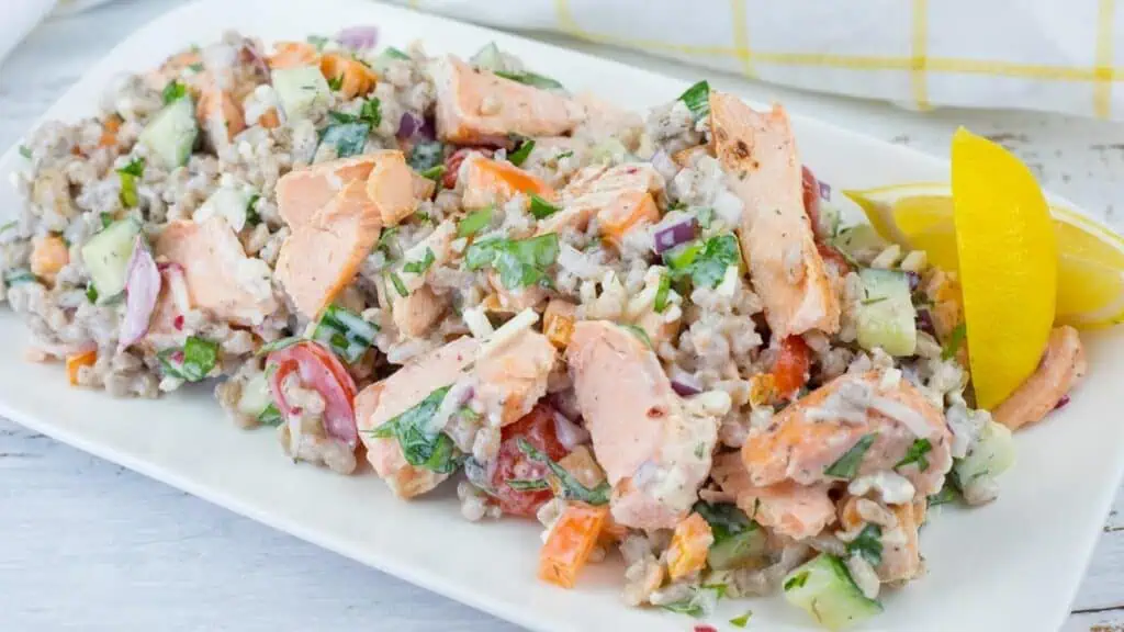 A plate of mixed salad with chunks of salmon, diced vegetables, grains, and herbs, garnished with two lemon wedges.