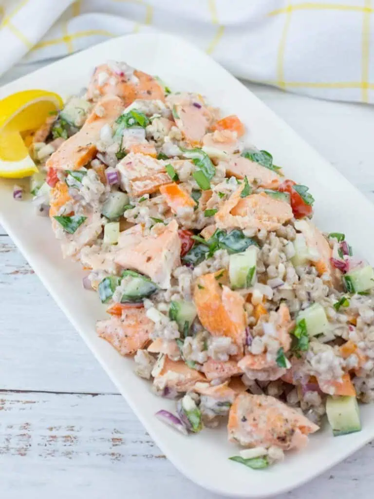 A rectangular white plate with a barley salad containing flaked salmon, cucumbers, red onions, and herbs, garnished with two lemon wedges.