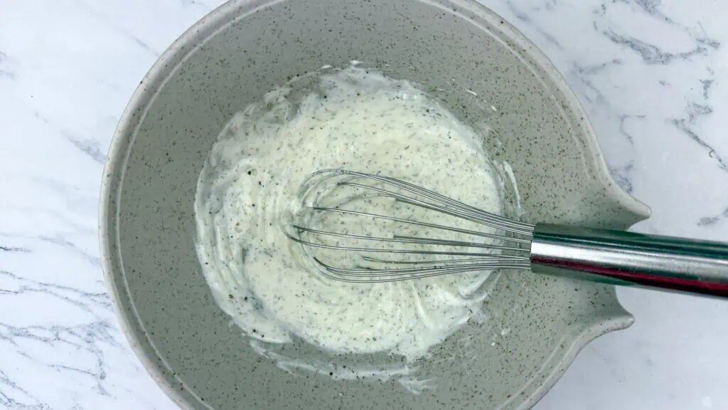A metal whisk rests in a speckled mixing bowl containing a creamy white sauce or dressing on a marble countertop.