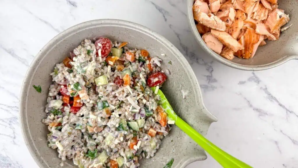 A bowl of mixed vegetable salad with grains and a creamy dressing next to a smaller bowl of flaked cooked salmon on a white marble surface.