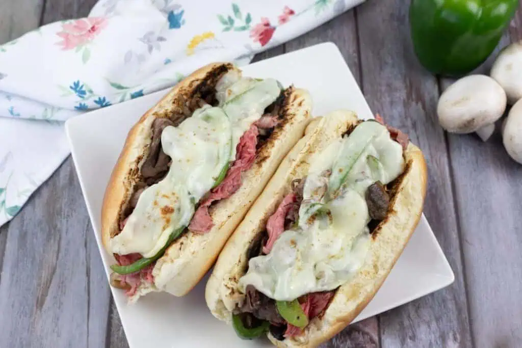 Two cheesesteak sandwiches topped with melted cheese, sliced beef, green peppers, and mushrooms on a white square plate.