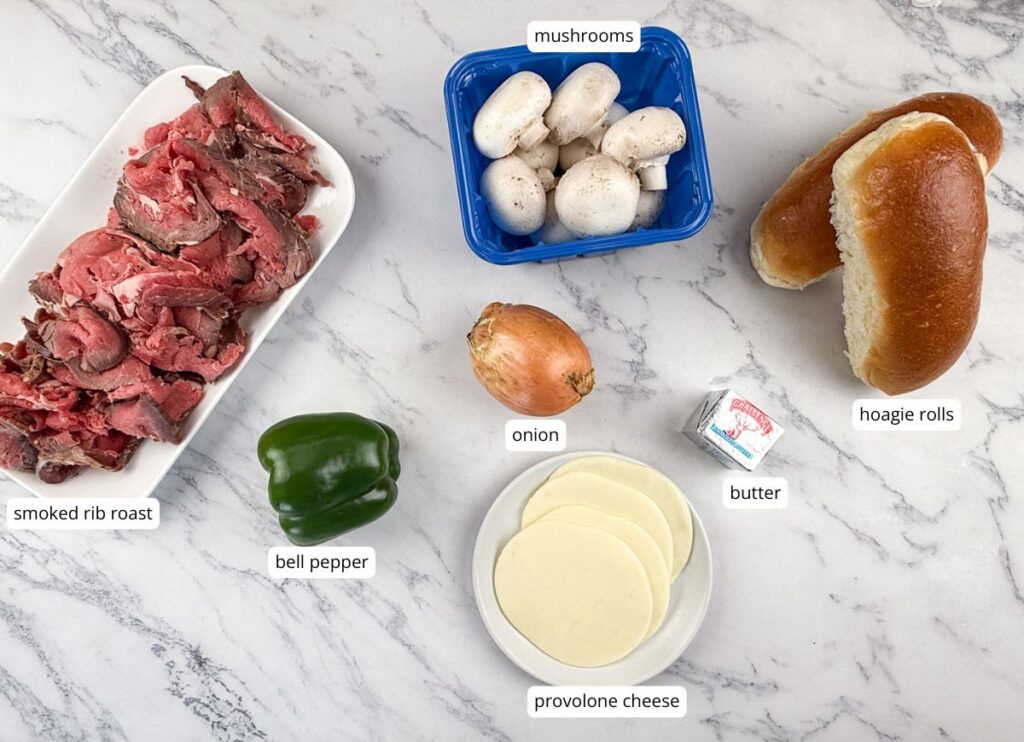 Ingredients for a sandwich laid out on a marble surface: smoked rib roast, green bell pepper, onion, mushrooms, butter, provolone cheese, and hoagie rolls, each labeled.