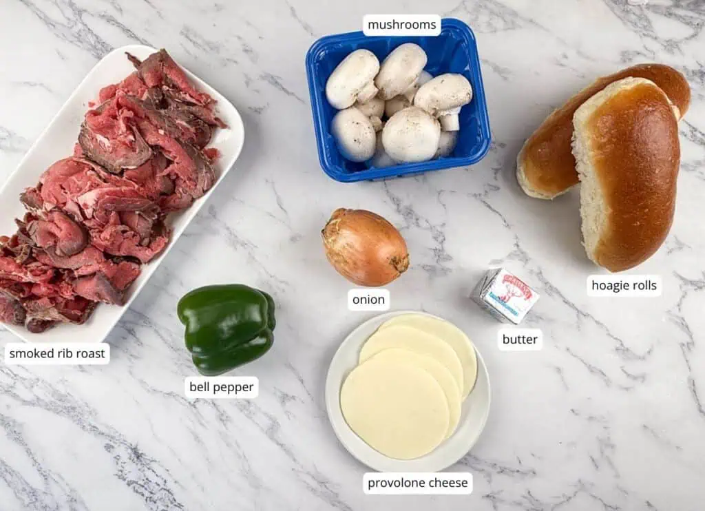 Ingredients for a sandwich laid out on a marble surface: smoked rib roast, green bell pepper, onion, mushrooms, butter, provolone cheese, and hoagie rolls, each labeled.