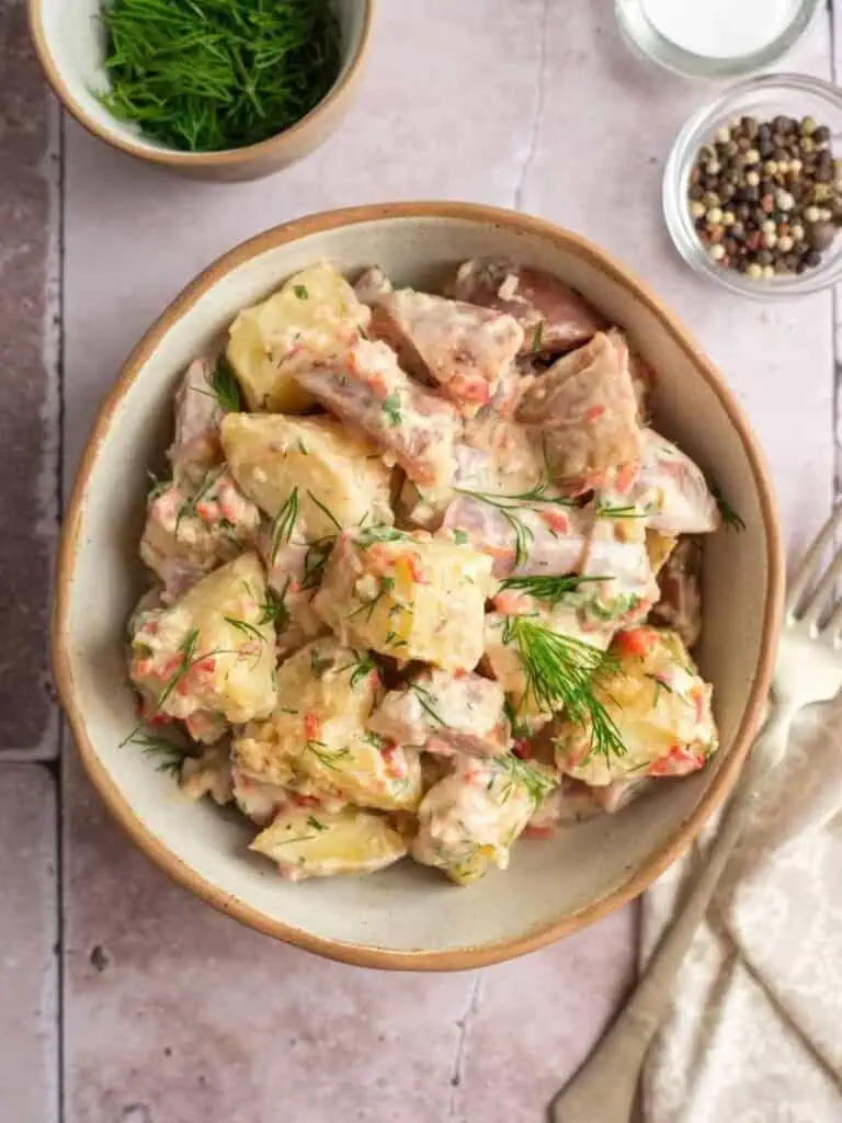 A bowl of potato salad with dill, carrot pieces, and creamy dressing, placed on a tiled surface next to a small bowl of fresh dill and dishes of salt and pepper.