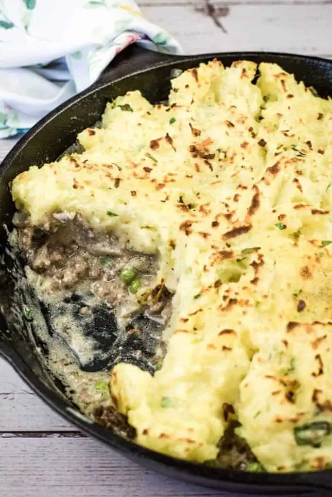 A cast-iron skillet filled with stroganoff pie, showing a layer of browned mashed potatoes over ground meat and vegetables with a serving removed.