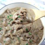 A wooden spoon holds a serving of beef stroganoff with mushrooms and creamy sauce above a pot filled with the dish.