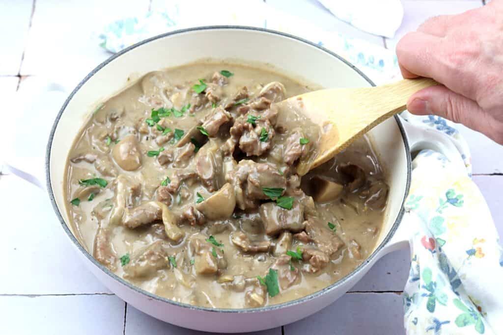 A hand stirs a pot of creamy beef and mushroom stew with a wooden spoon, garnished with chopped parsley.