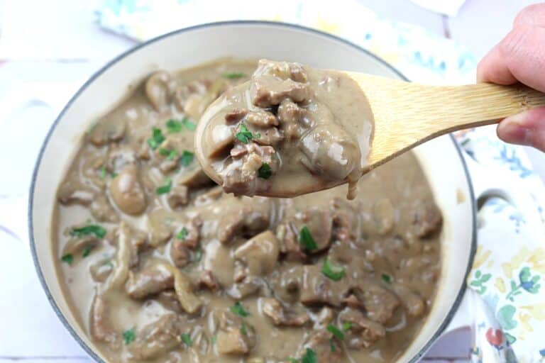 A wooden spoon holds a serving of beef stroganoff with mushrooms and creamy sauce above a pot filled with the dish.