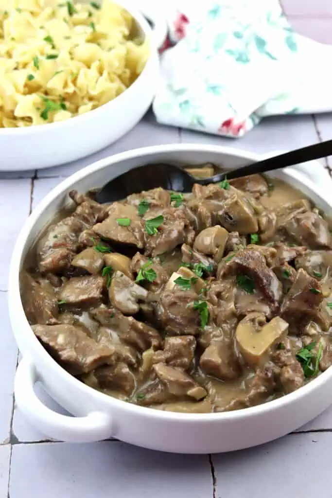A white bowl of beef stroganoff with mushrooms and sauce, garnished with parsley, sits next to a bowl of cooked egg noodles.