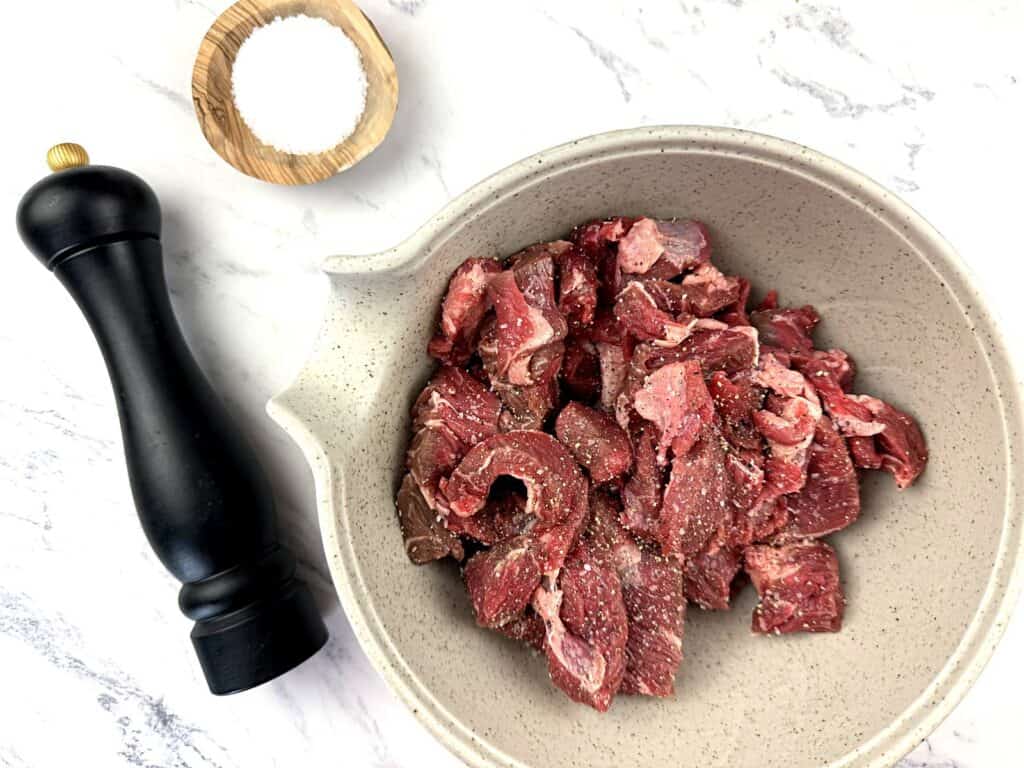 A bowl of raw sliced beef seasoned with salt and pepper, next to a wooden bowl of salt and a black pepper grinder on a white marble surface.