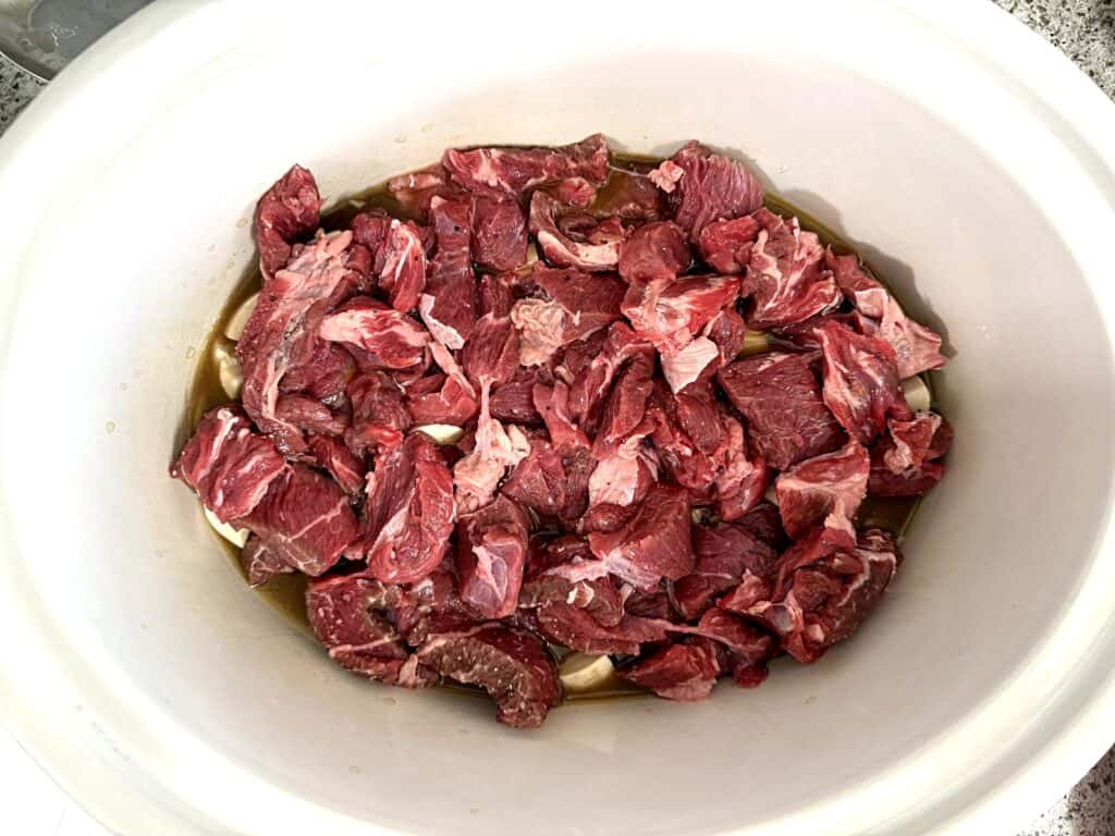 Raw beef chunks in a white slow cooker, partially submerged in liquid, ready for cooking.