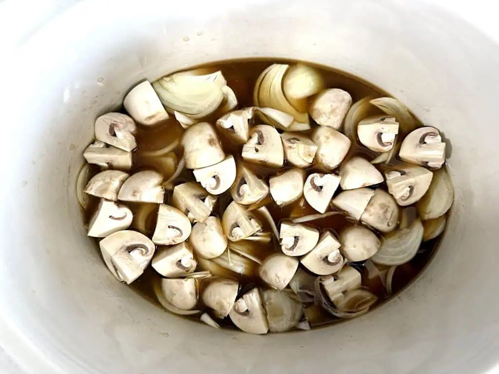 Chopped mushrooms and onion slices in a brown liquid inside a white slow cooker or pot.