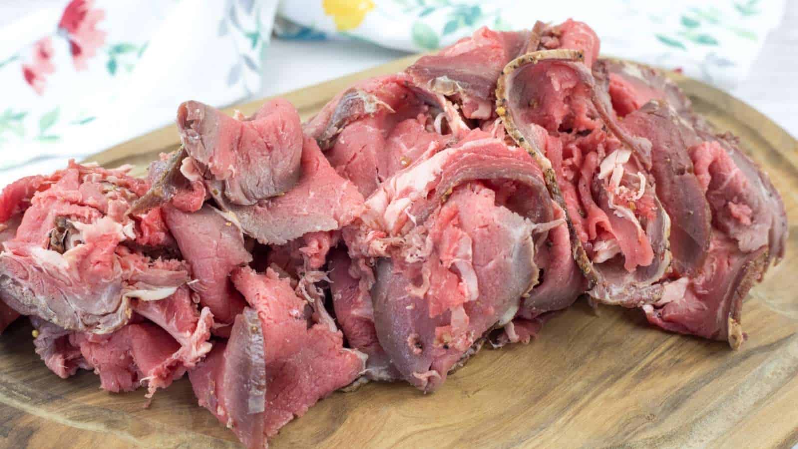 Thin slices of rare roast beef piled on a wooden cutting board with a floral-patterned cloth in the background.