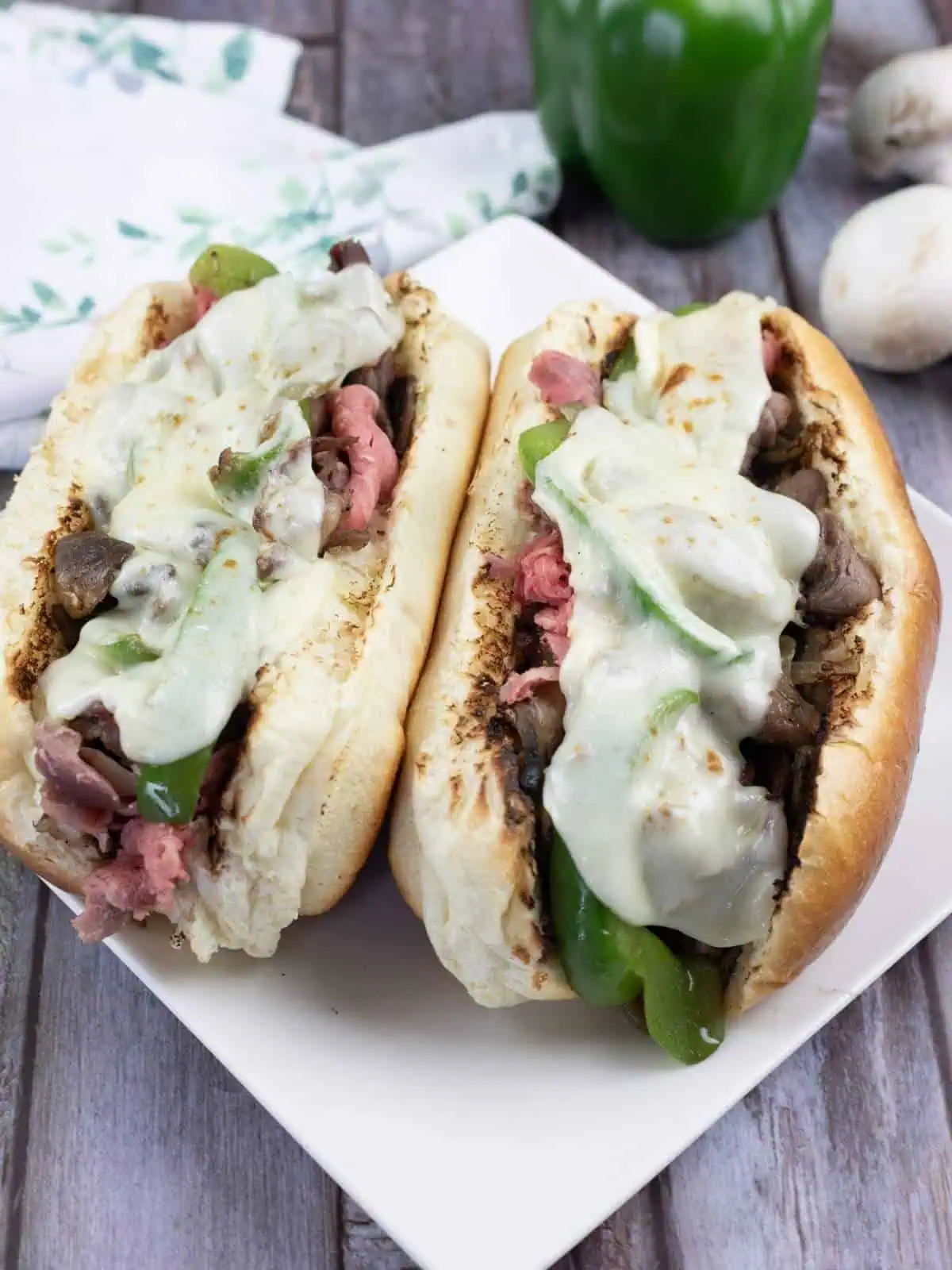 Two Philly cheesesteak sandwiches with sliced beef, green bell peppers, mushrooms, and melted cheese on hoagie rolls, served on a white plate.