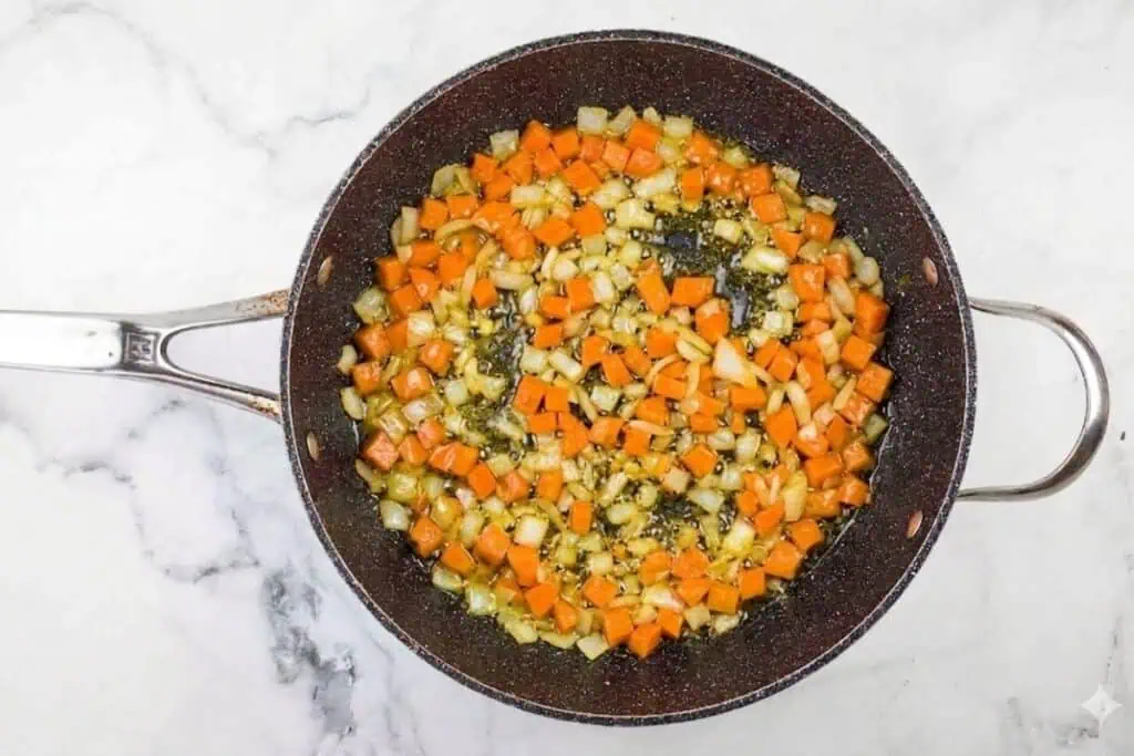 Diced carrots, celery, and onions are sautéing in oil in a large skillet on a white marble surface.