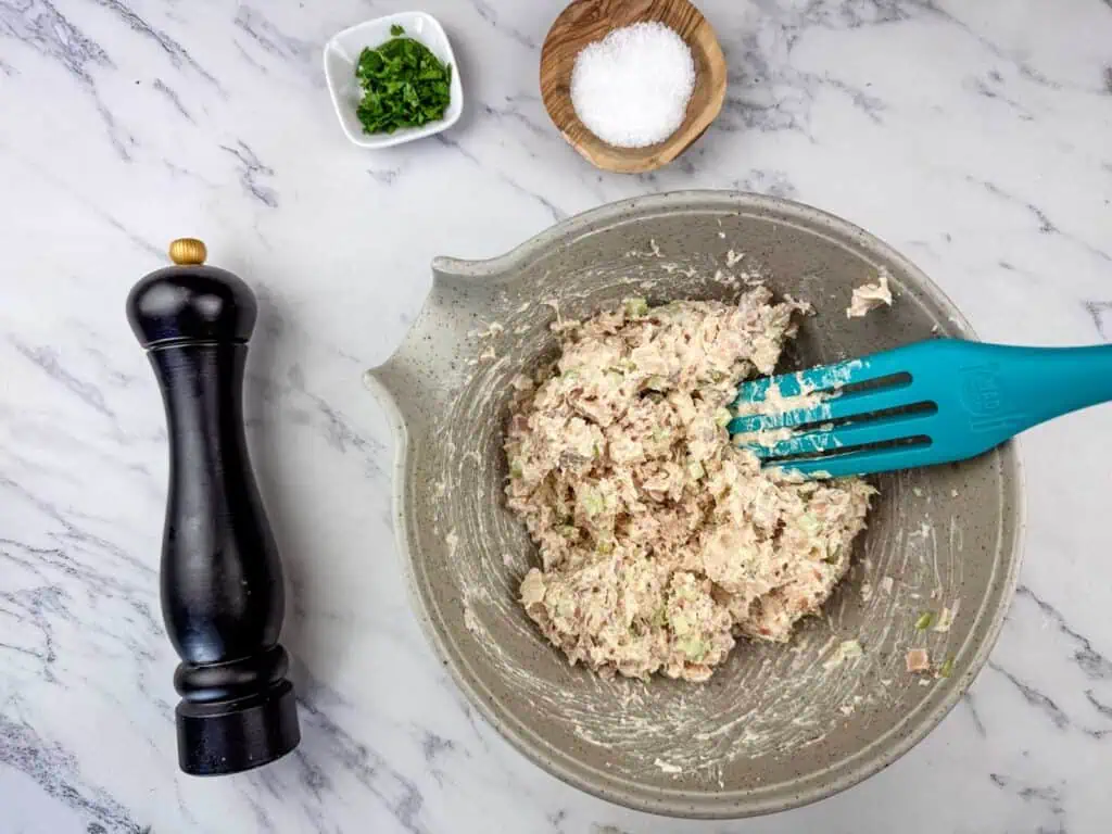 A bowl of tuna salad being mixed with a spatula, surrounded by a pepper grinder, a small bowl of chopped herbs, and a bowl of salt on a marble countertop.