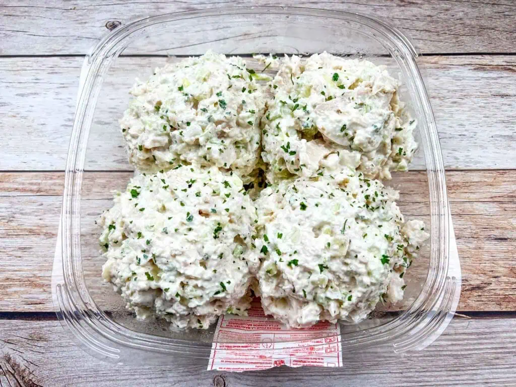 Plastic container holding four scoops of chicken salad, garnished with chopped herbs, on a wooden surface.