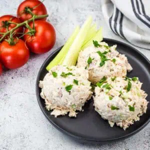 A black plate with three scoops of Costco chicken salad garnished with parsley and celery sticks, next to a cluster of vine tomatoes.