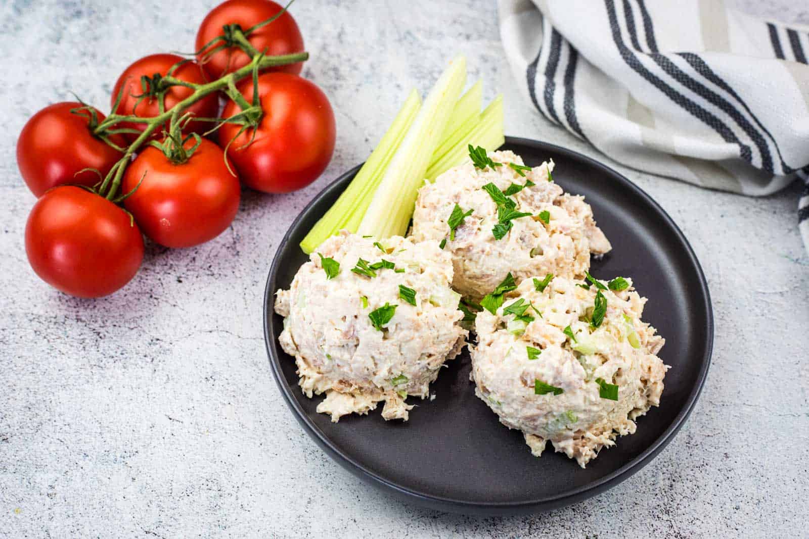 A black plate with three scoops of Costco chicken salad garnished with parsley and celery sticks, next to a cluster of vine tomatoes.