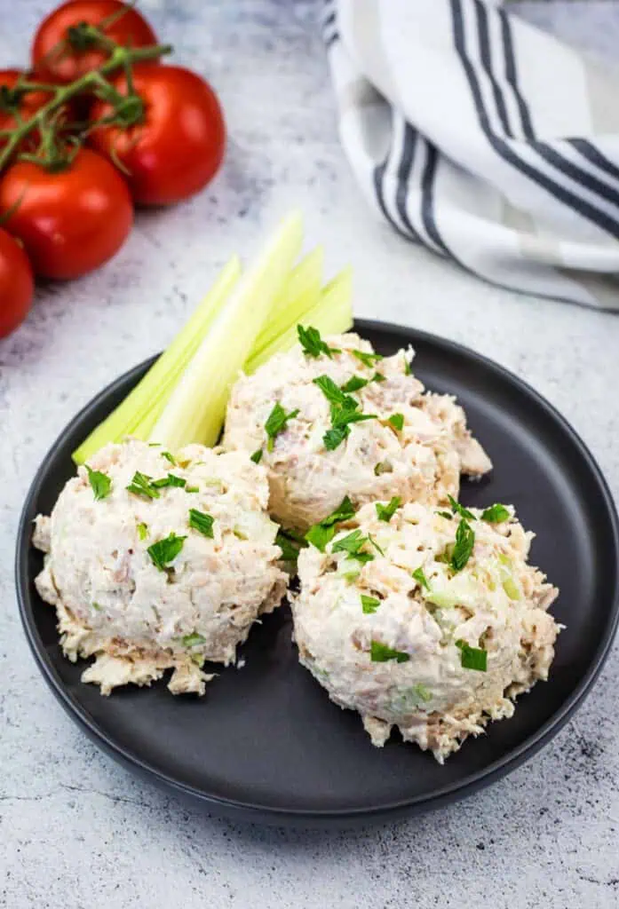 Three scoops of tuna salad garnished with chopped herbs are served on a black plate with celery sticks; tomatoes and a striped towel are in the background.