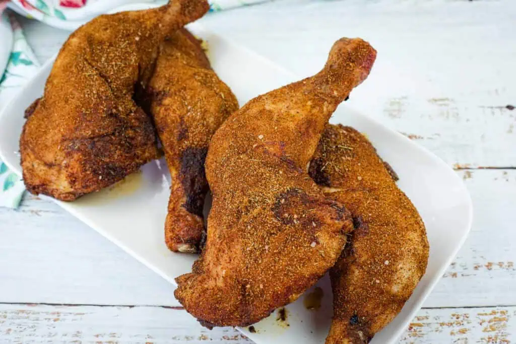 Three seasoned and cooked chicken legs are arranged on a white rectangular plate, placed on a rustic white wooden surface.
