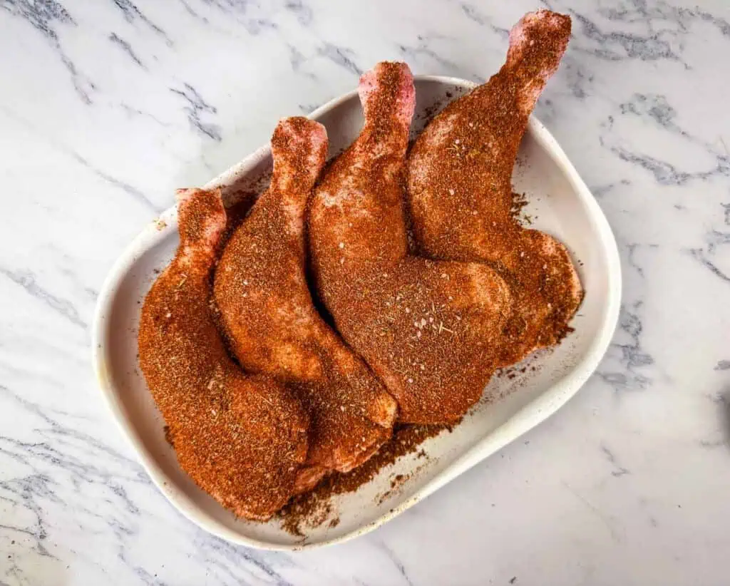 Four raw chicken leg quarters coated with a reddish spice rub are arranged on a white oval dish on a marble surface.