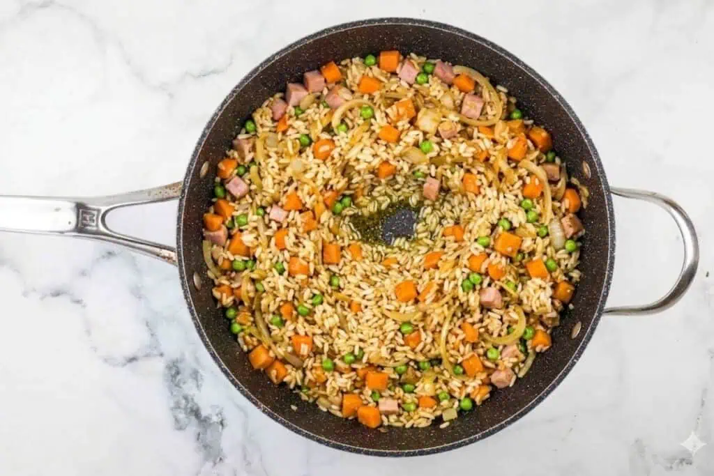 A pan of fried rice with diced ham, peas, carrots, and onions on a marble countertop.