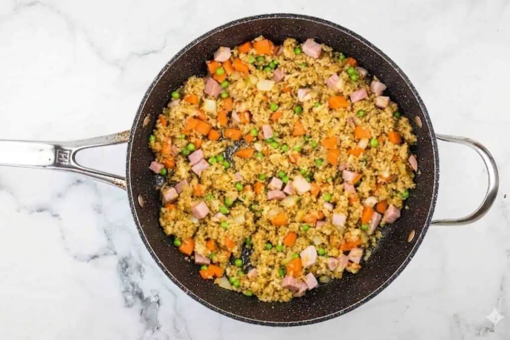 A large black skillet filled with fried rice, diced ham, carrots, peas, and egg, sitting on a white marble surface.