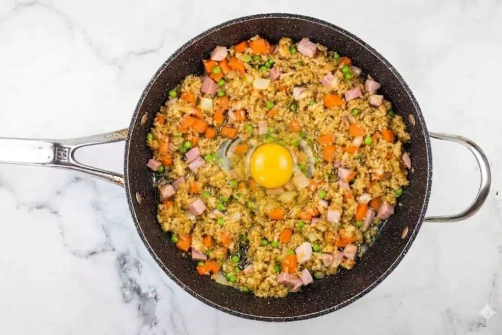 A frying pan with cooked rice, diced ham, carrots, peas, and an uncooked egg in the center on a marble countertop.