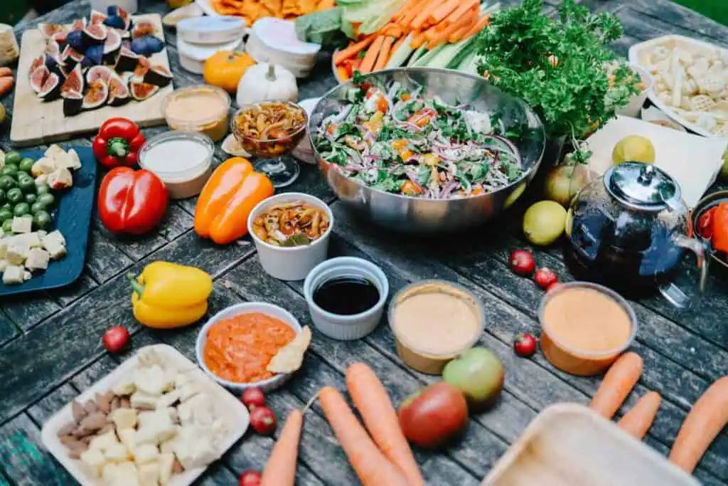 A wooden table covered with various fresh vegetables, dips, salads, cheese, fruits, and a teapot, arranged for a picnic or outdoor meal.