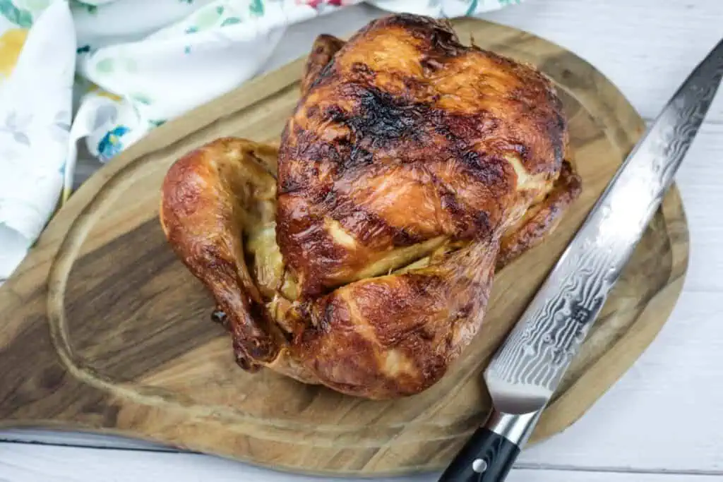 A whole roasted chicken sits on a wooden cutting board next to a carving knife.
