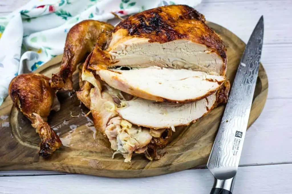 A roasted, partially carved chicken with sliced breast meat and a detached drumstick sits on a wooden cutting board beside a large knife.