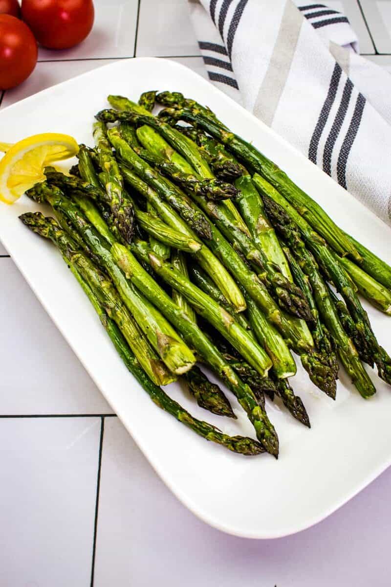 A white rectangular plate of smoked asparagus garnished with lemon slice, set on a tiled surface with a striped kitchen towel and tomatoes in the background.