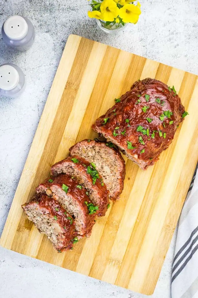 Sliced smoked meatloaf garnished with herbs on a wooden cutting board, alongside salt and pepper shakers.