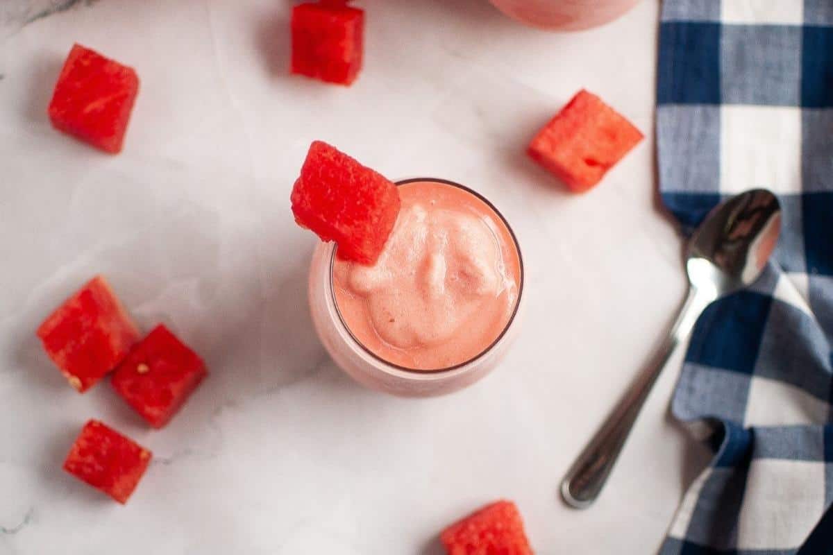 A glass of watermelon smoothie topped with a watermelon cube, surrounded by watermelon pieces, a spoon, and a blue checkered cloth on a white surface.