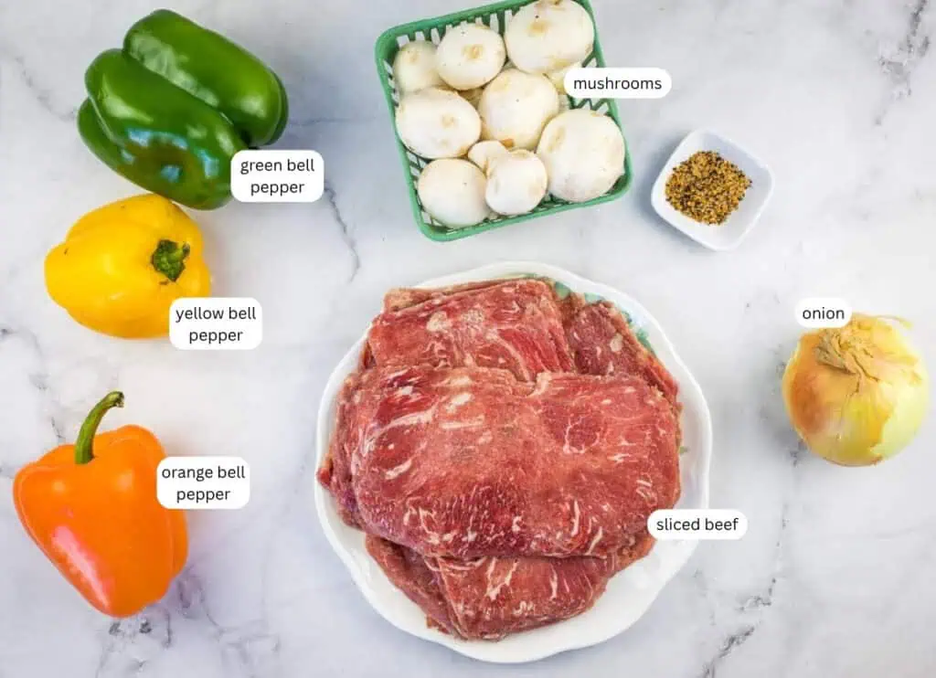Plate of sliced beef, whole green, yellow, and orange bell peppers, a basket of mushrooms, a whole onion, and a small dish of seasoning on a white countertop.