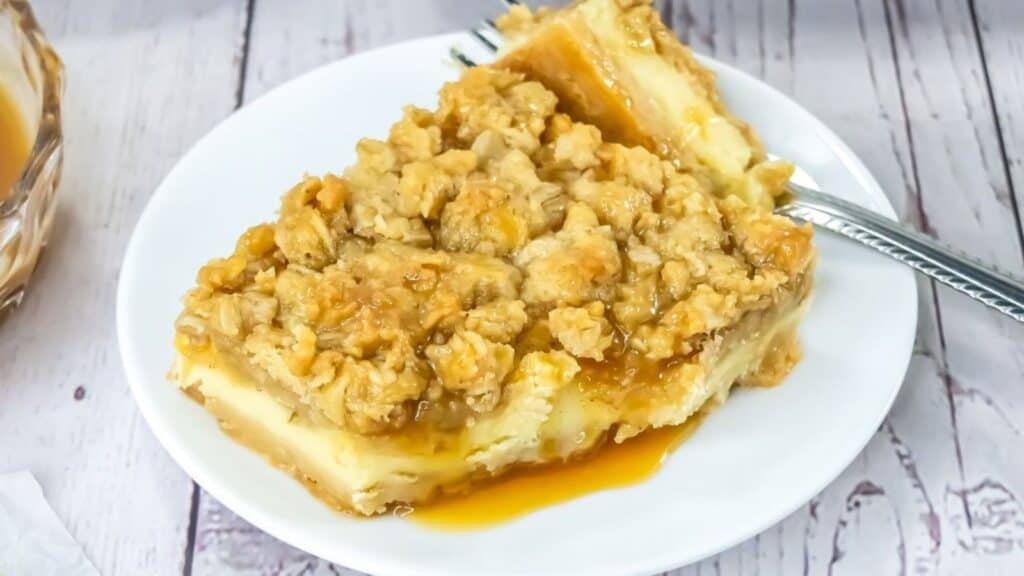 A square slice of crumb-topped dessert with caramel drizzle is served on a white plate with a fork.