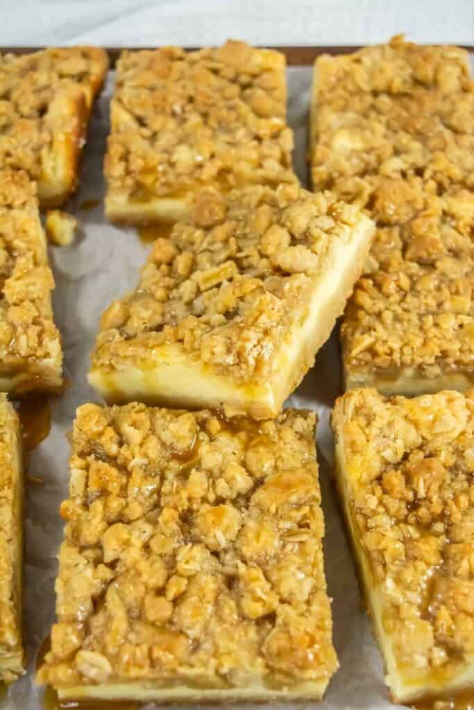 Several rectangular cheesecake bars with a crumbly oat topping and caramel drizzle are arranged on parchment paper.