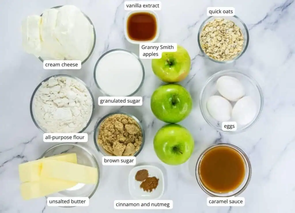 Top-down view of ingredients in bowls and cups, including cream cheese, oats, flour, sugars, eggs, butter, apples, vanilla extract, caramel sauce, and spices on a marble surface.